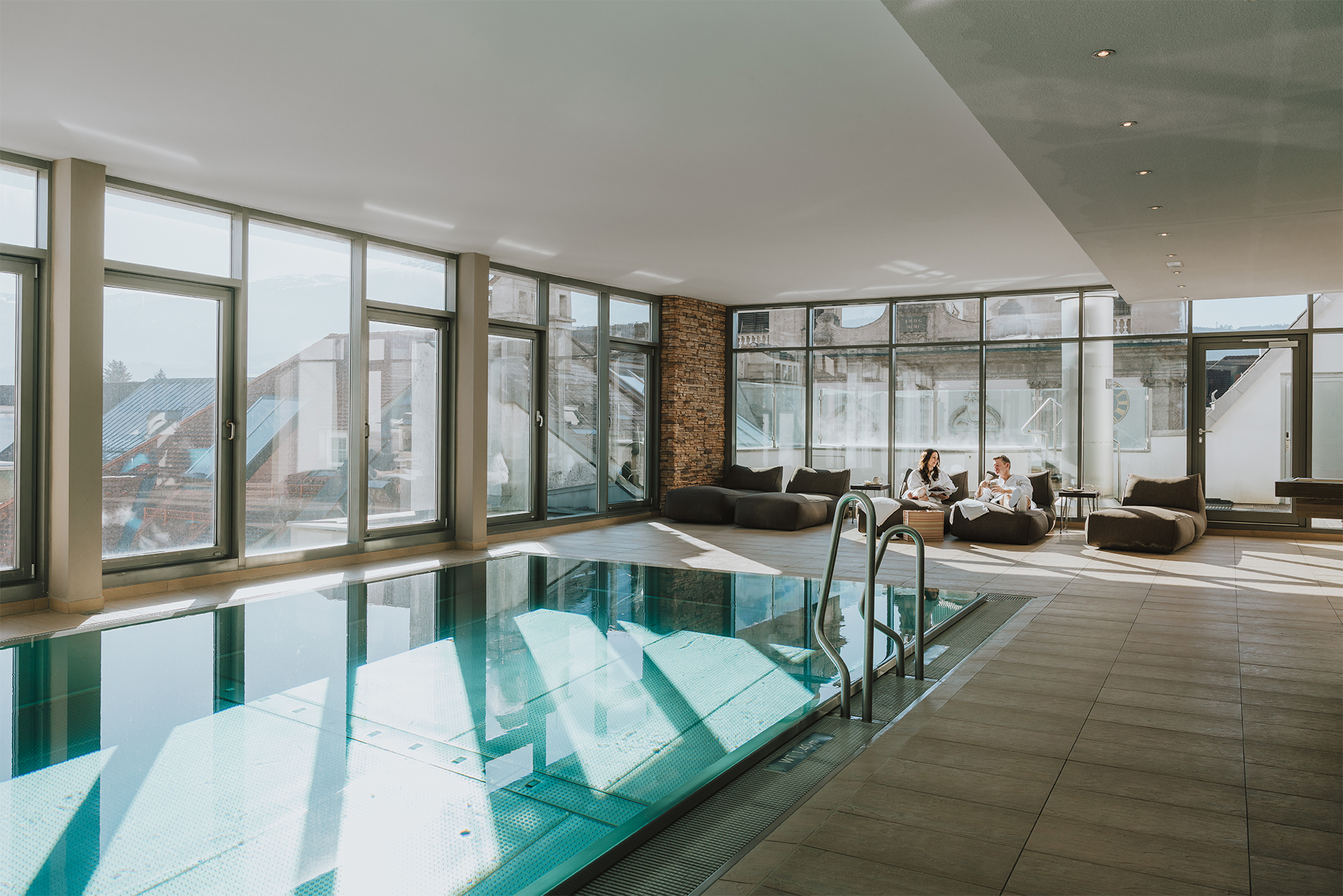 20250319-GrauerBaer_ohne_Grain-_N3A8793-©Roman-Huber Innenpool im Rooftop Wellnessbereich im Hotel Grauer Bär Innsbruck mit großen Fenstern und zwei Gästen auf Sitzpolstern entspannend.